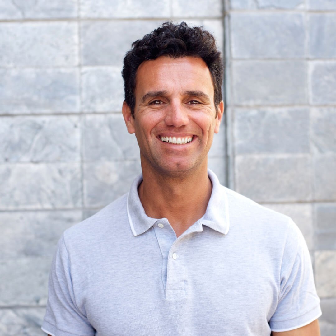 Smiling man with dark hair wearing a light gray polo shirt against a gray tiled wall