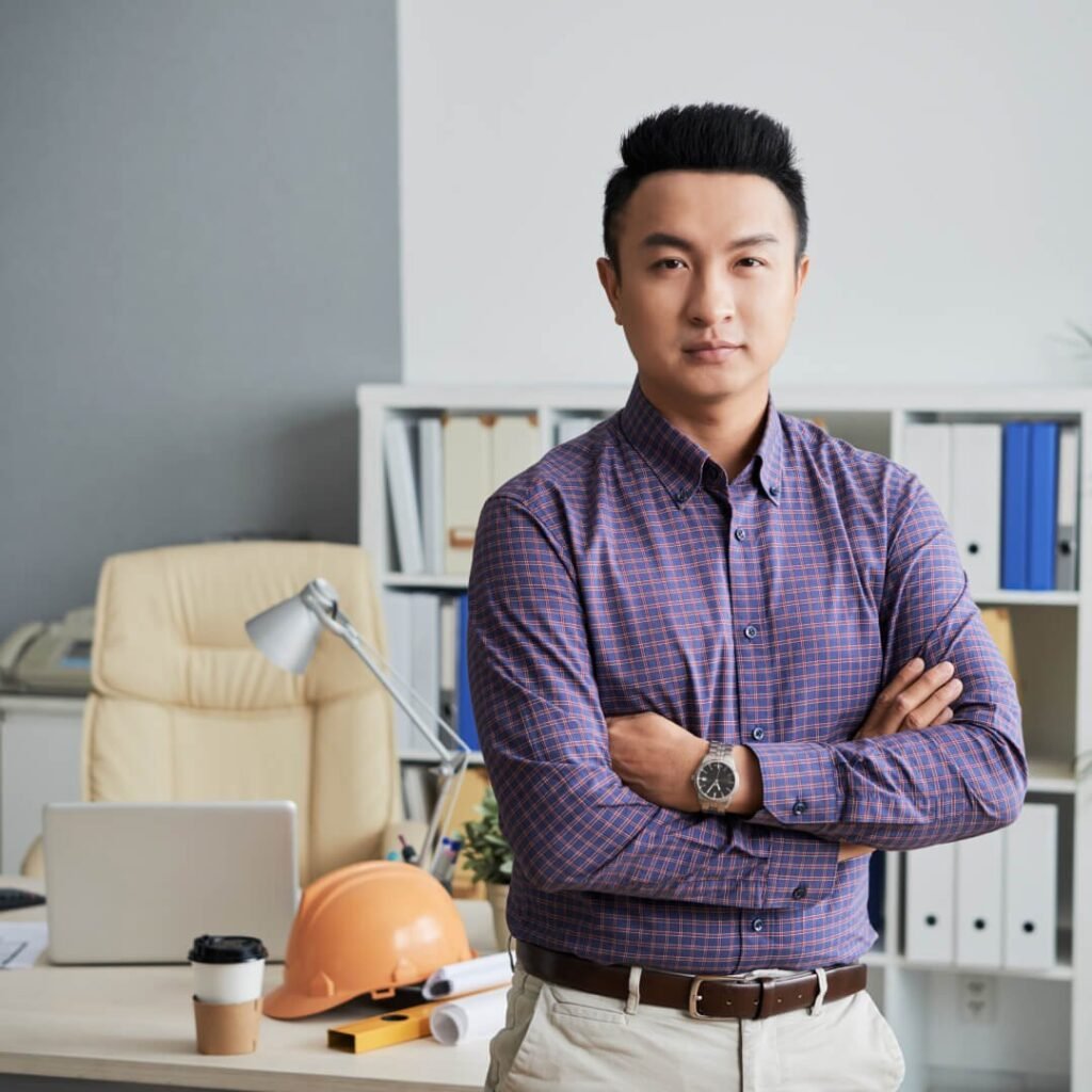 Professional man in a plaid shirt standing with arms crossed in an office with a hard hat on his desk