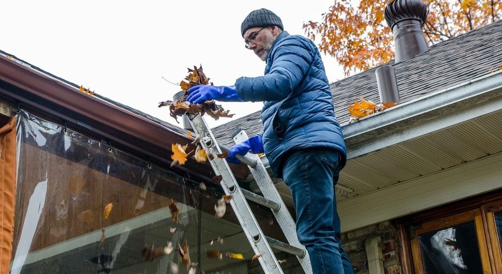 Man on a ladder wearing blue gloves clearing autumn leaves from a house gutter